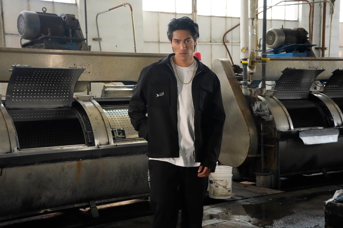 Person standing in a factory setting with large industrial machines wearing the LLC Plaque Zip Jacket.