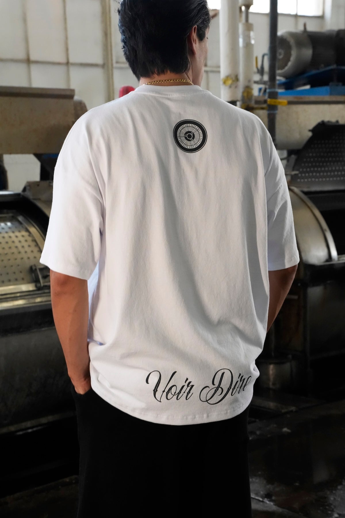 Back of model wearing a Heritage t-shirt with Voir Dire text at the hem standing in an industrial setting (White)