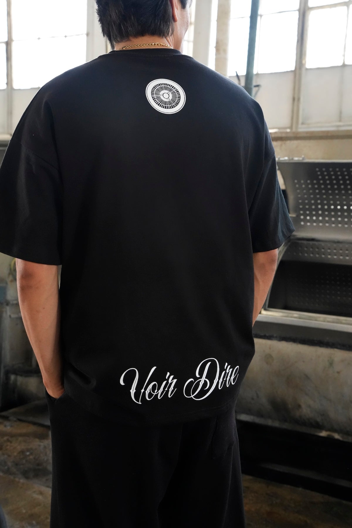 Model wearing the Heritage black t-shirt with 'Voir Dire' text on the back, standing in an industrial setting. (Black)