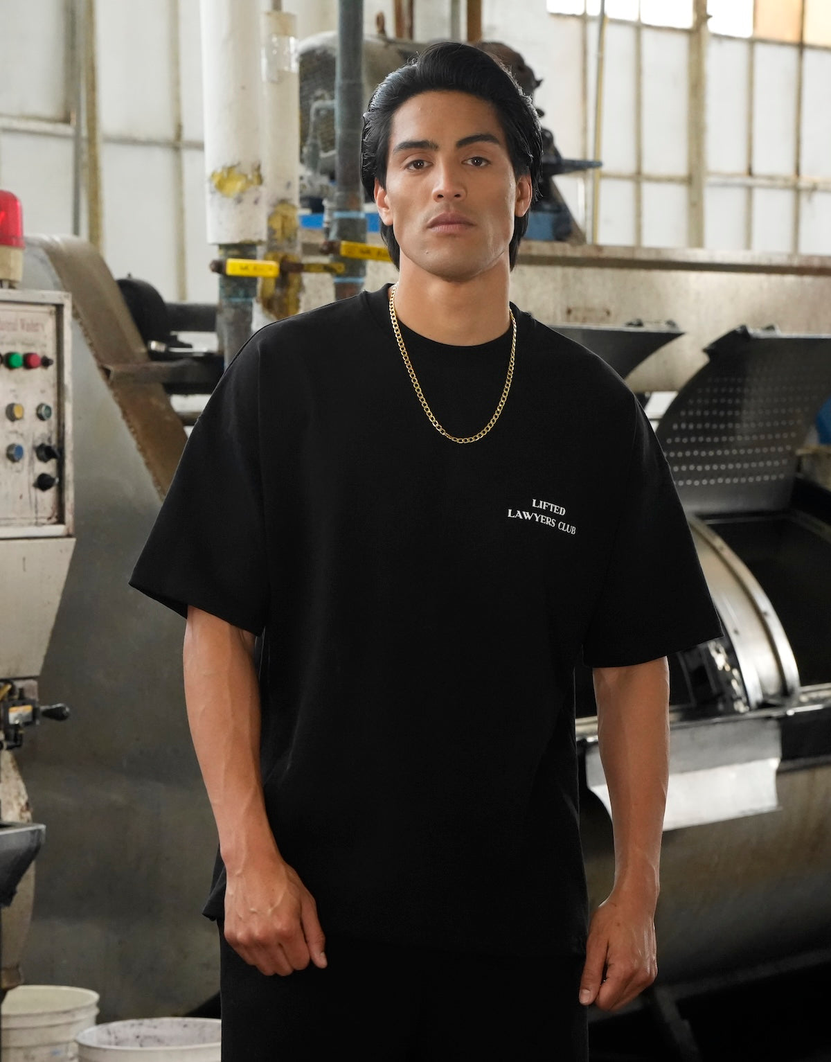 Model wearing the Heritage logo black t-shirt with text in an industrial setting (Black)